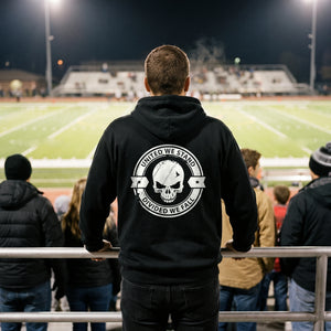 Man in the black United We Stand Hoodie at a hometown Friday night football game, seen from behind with the white skull seal across the back
