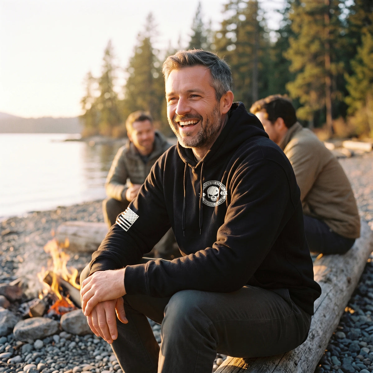 Man in the black United We Stand Hoodie laughing with friends by a lakeside campfire, white skull seal on the left chest and flag on the right sleeve