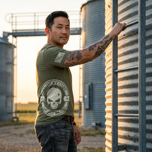 Man in right-side profile beside a grain bin at a farm co-op at golden hour, one hand on the steel ladder rail, wearing the United We Stand Skull T-Shirt in Military Green: United We Stand, Divided We Fall skull seal printed around the right side at the hip, American flag on the right sleeve