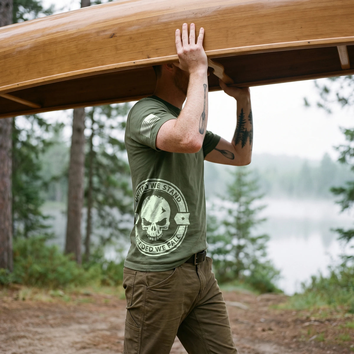 Man carrying a wooden canoe overhead on a forest lake trail, seen from his right side, wearing the United We Stand Skull T-Shirt in Military Green: United We Stand, Divided We Fall skull seal printed around the right side at the hip, American flag on the right sleeve