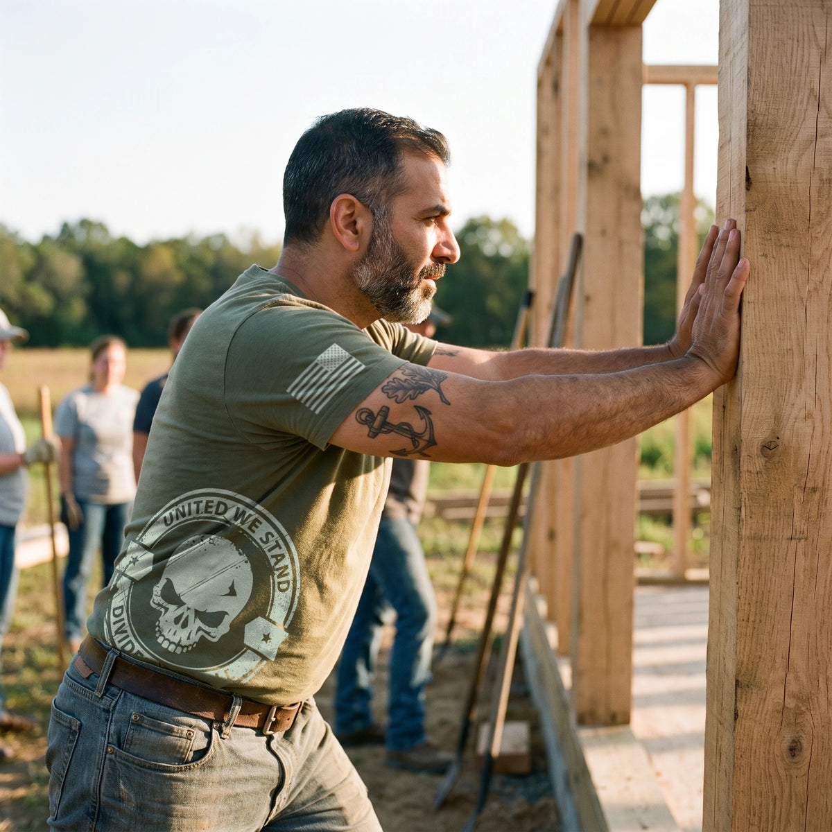 Man helping a volunteer crew raise a timber frame, pushing a wooden post with both hands, wearing the United We Stand Skull T-Shirt in Military Green: United We Stand, Divided We Fall skull seal printed around the right side at the hip, American flag on the right sleeve