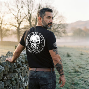 Man by a dry-stone wall in a misty meadow at dawn, looking back over his shoulder, wearing the We the People T-Shirt in Black: Liberty or Death skull seal printed large on the back, American flag on the right sleeve
